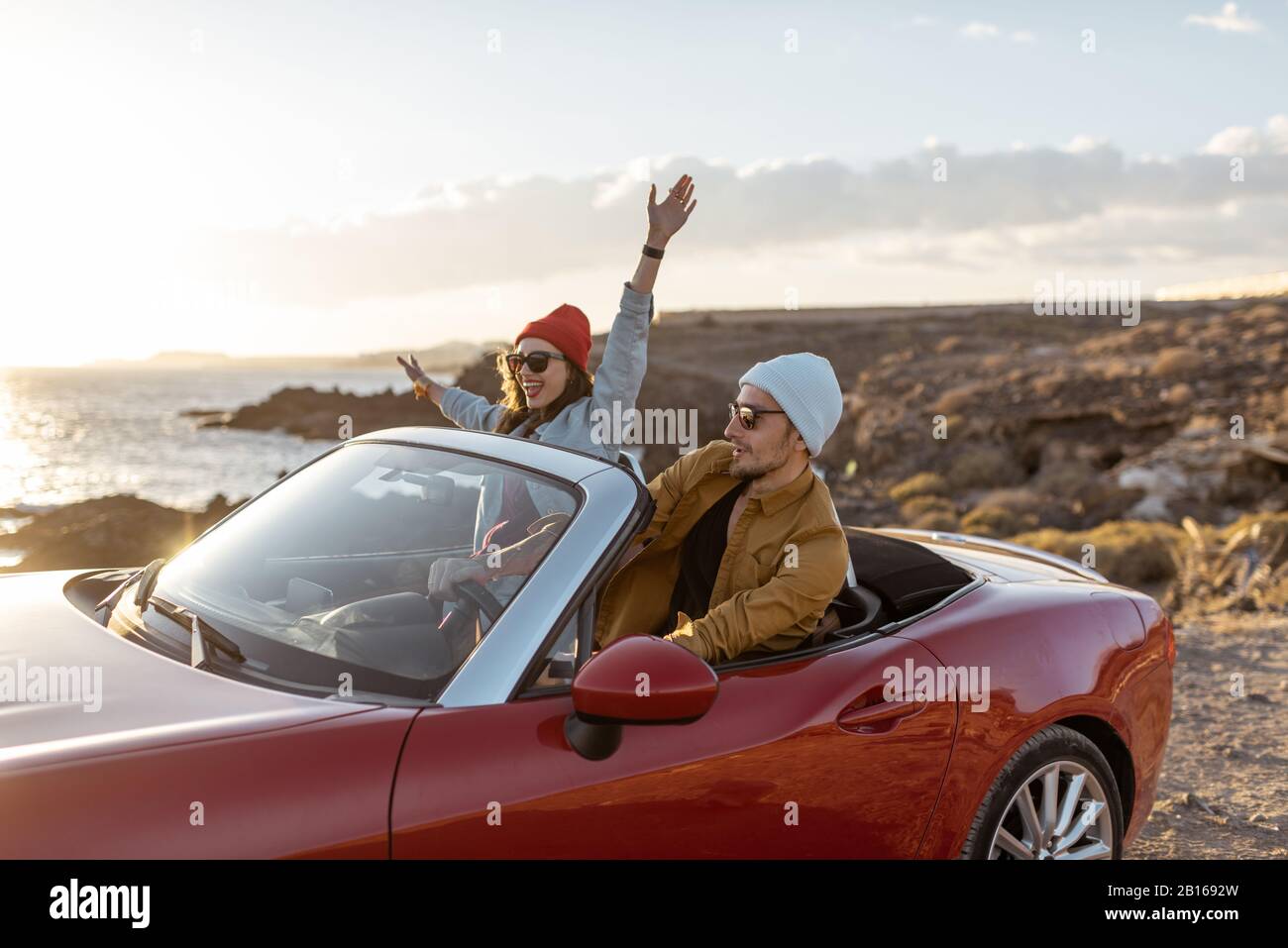 Joyful couple enjoying vacations, driving together convertible car on the rocky ocean coast on a sunset. Happy vacation, love and travel concept Stock Photo