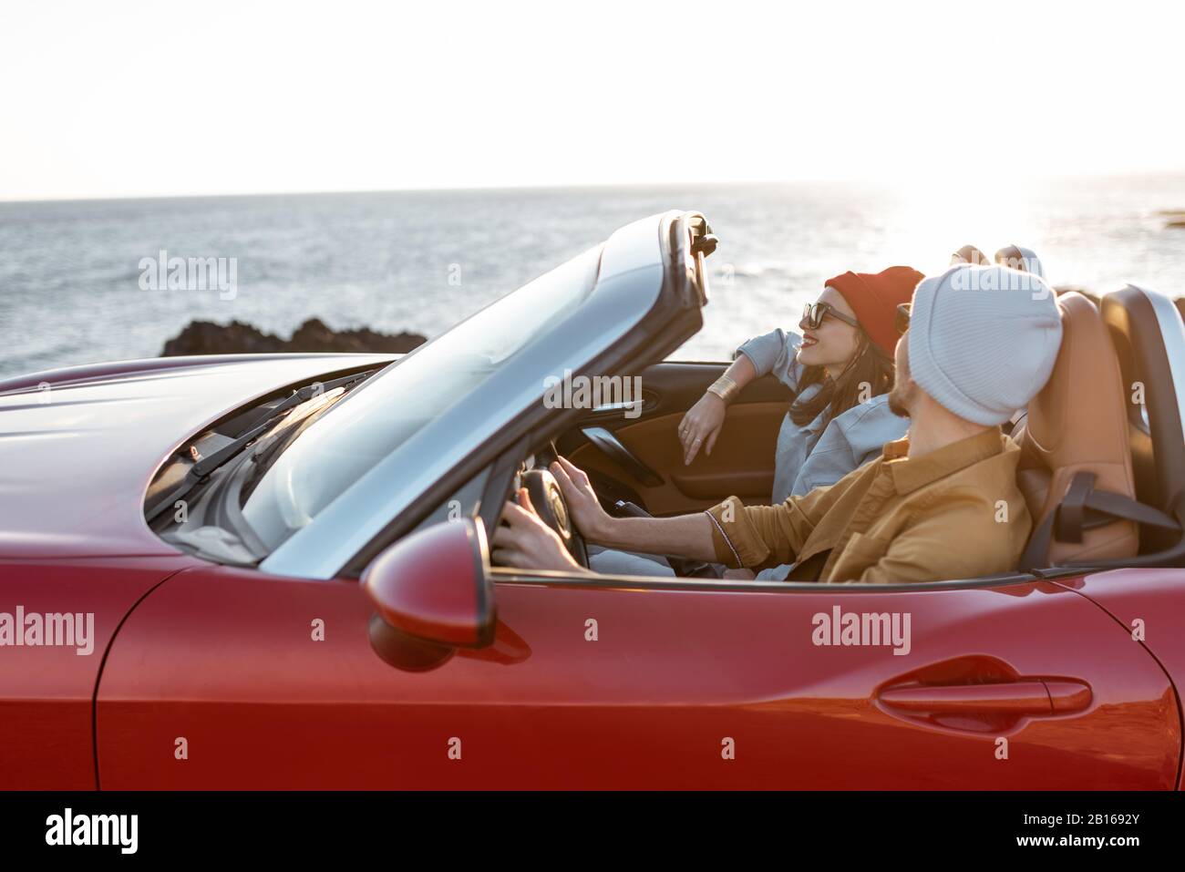 Joyful couple enjoying vacations, driving together convertible car on the rocky ocean coast on a sunset. Happy vacation, love and travel concept Stock Photo