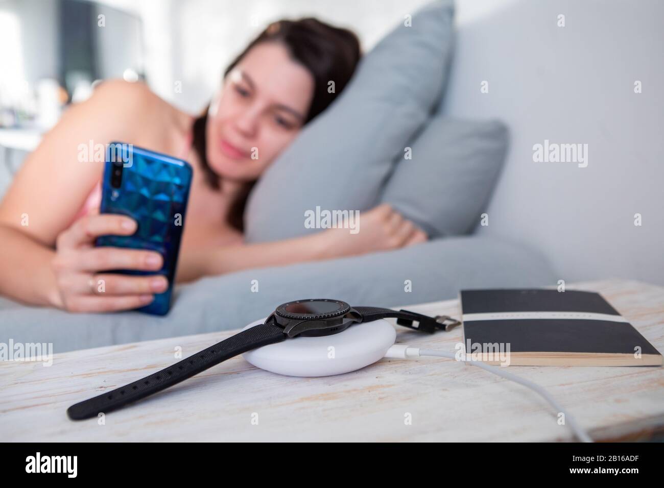 woman surfing internet on cellphone laying in bed smart watch on wireless charger Stock Photo