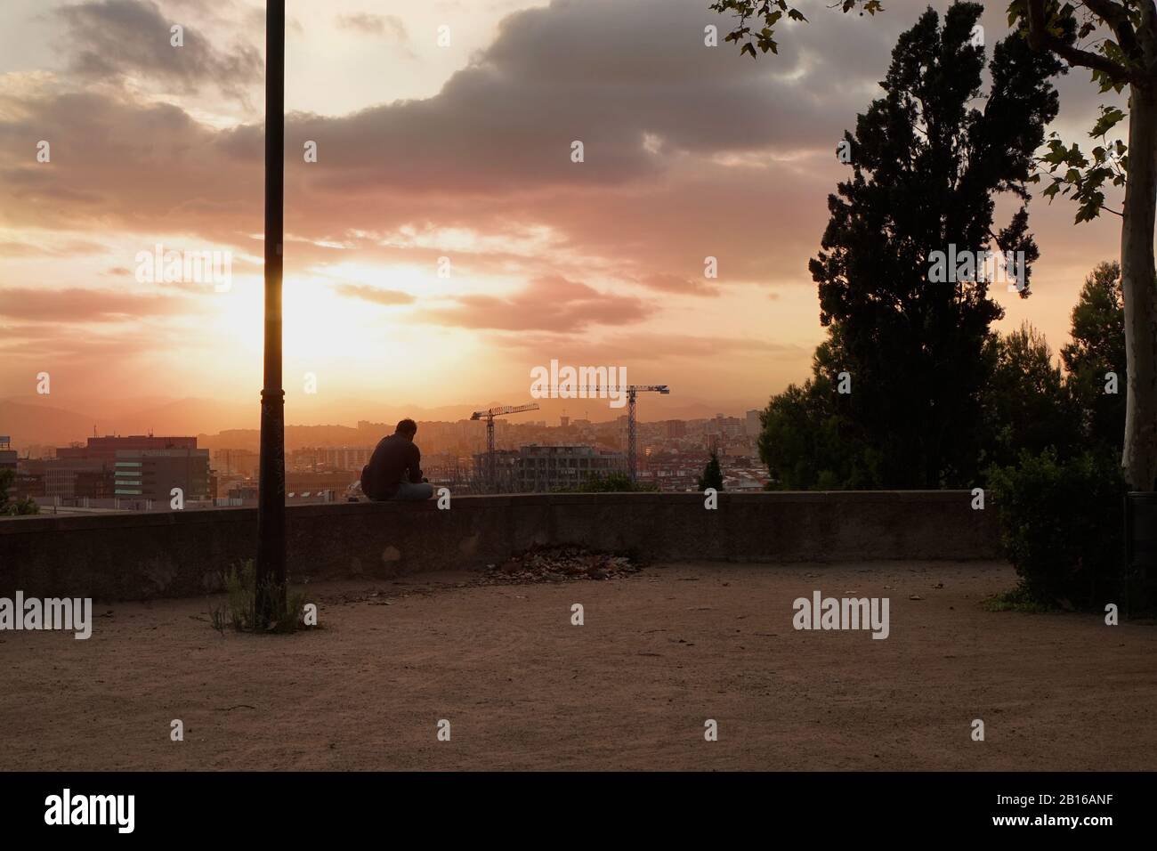 A man sits on a wall looking at the view of Barcelona outskirts city scape at twilight Stock Photo