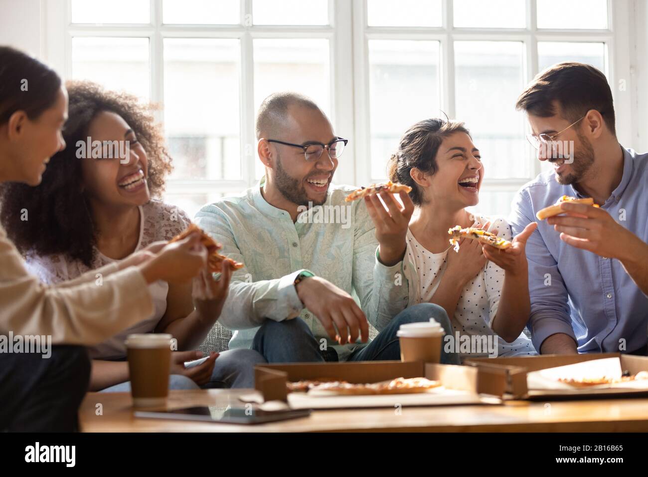 Five laughing multi-ethnic friends eating pizza indoors Stock Photo