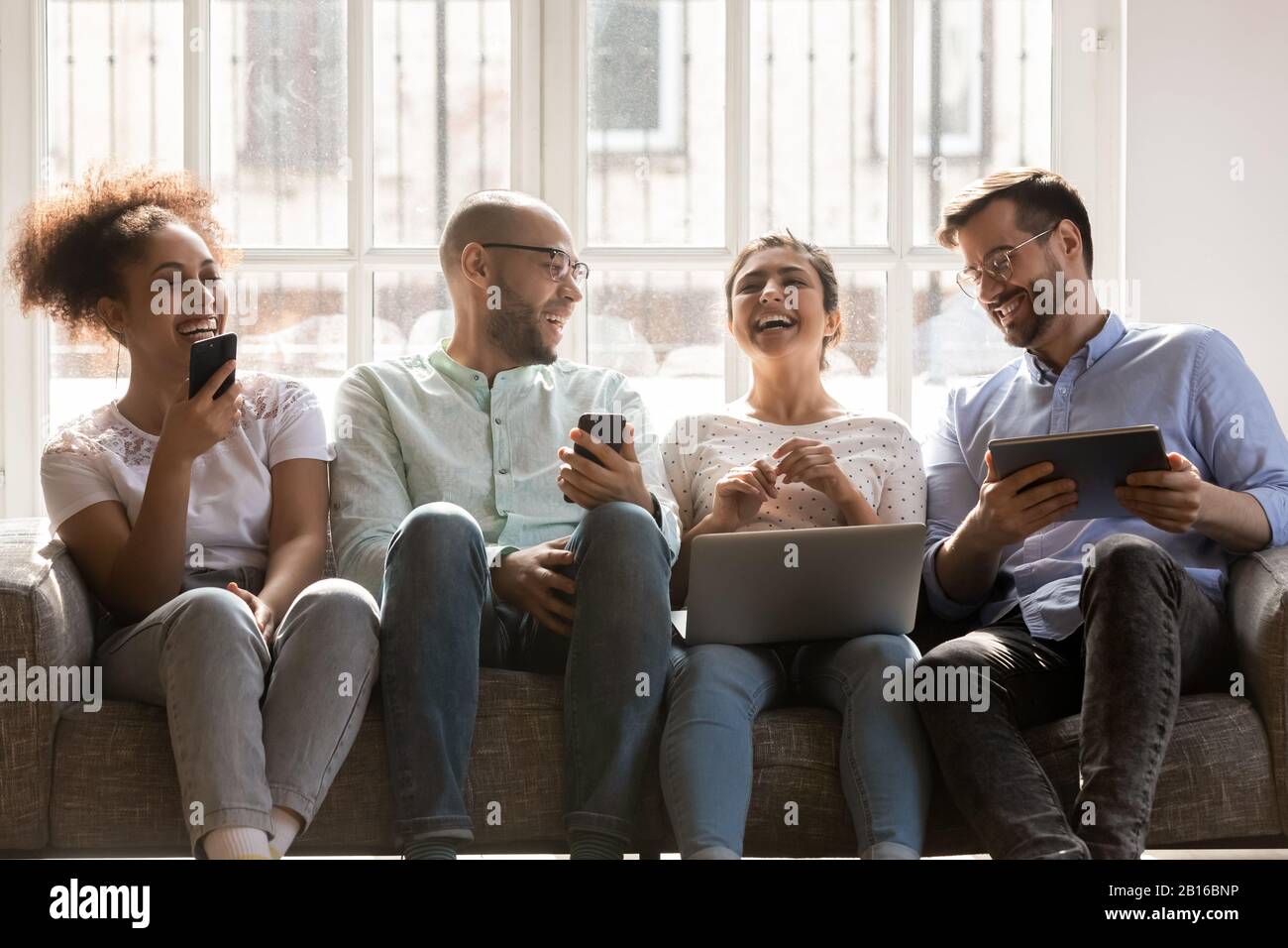 Cheerful multiracial friends using diverse gadgets sitting together on couch Stock Photo