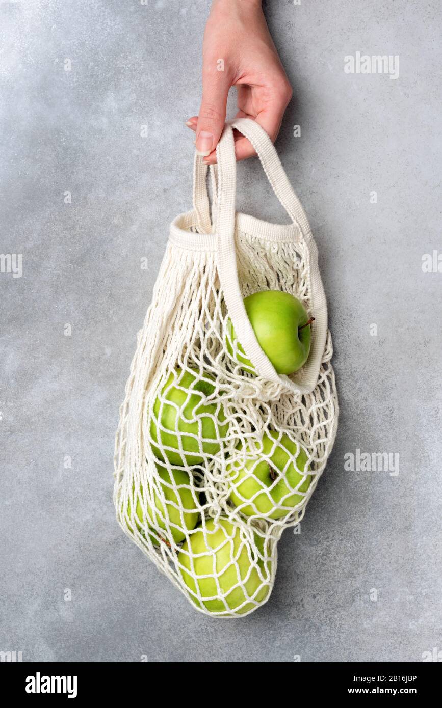 Hand holding mesh bag with green organic apples over grey concrete background. Concept of zero waste, reusable shopping bag, sustainable lifestyle Stock Photo