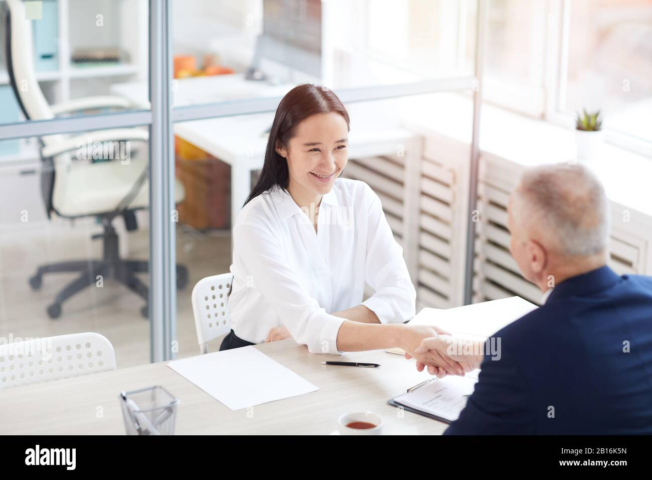 High angle portrait of young Asian businesswoman smiling happily while shaking hands with senior man across table in office, copy space Stock Photo