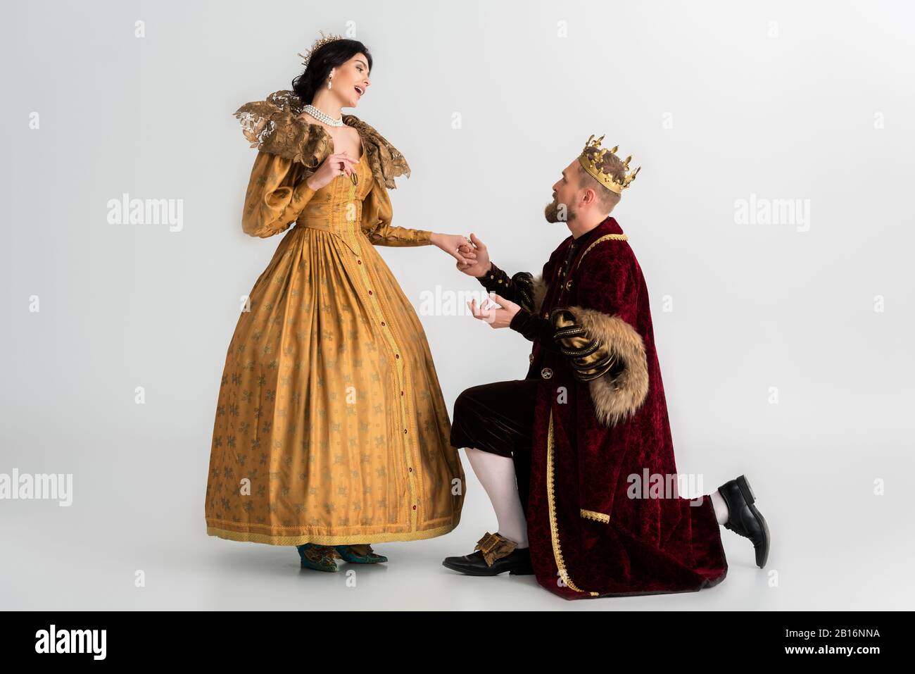 king with crown bending on knee and holding hand of smiling queen on grey  background Stock Photo - Alamy