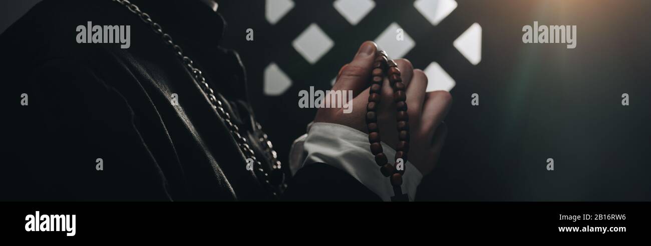 partial view of catholic priest holding wooden rosary beads near confessional grille in dark with rays of light, panoramic shot Stock Photo