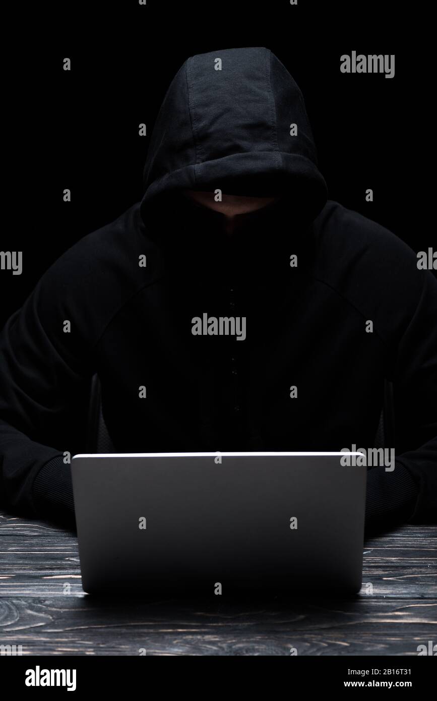 anonymous hacker in hood using laptop isolated on black Stock Photo