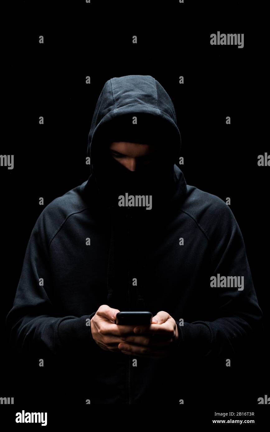 hacker in hood using smartphone isolated on black Stock Photo