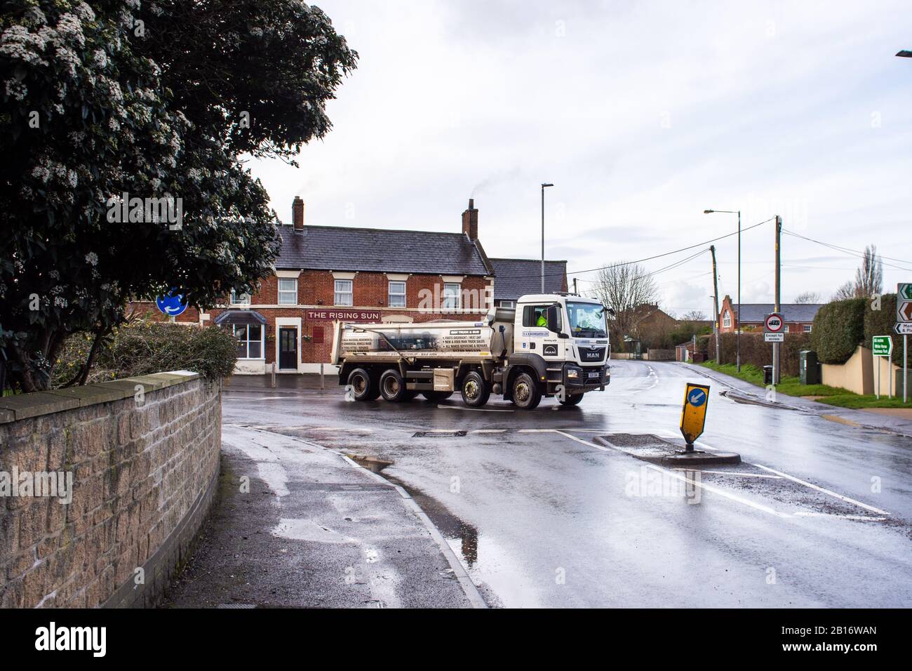 An eight wheeled MAN tipper lorry crossing a painted mini roundabout at a village junction after recent rain showers Stock Photo