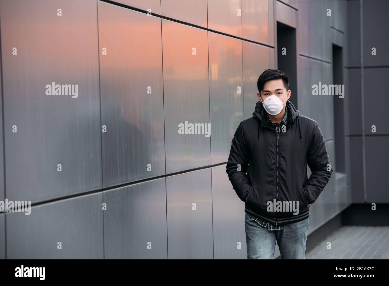 young asian man in respirator mask looking at camera while walking ...