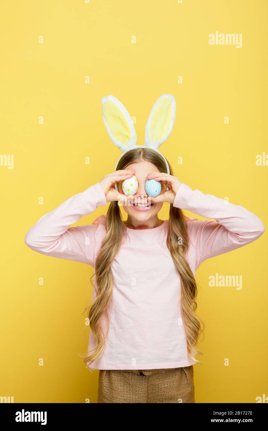 cheerful child with bunny ears covering eyes with easter eggs isolated on yellow Stock Photo