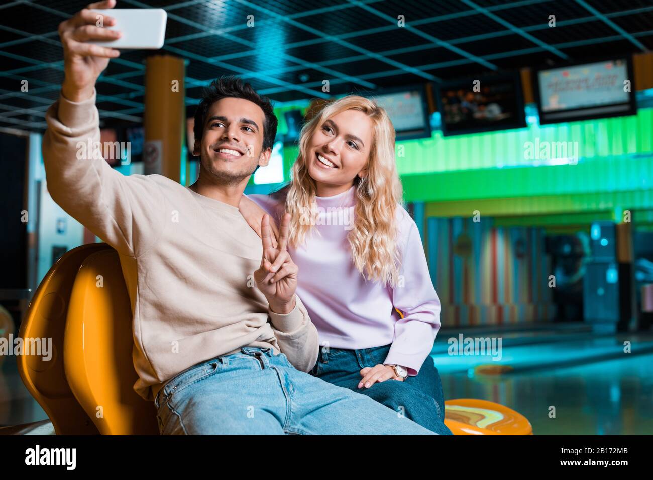happy young man showing victory gesture while sitting in bowilng club and taking selfie with smiling girlfriend Stock Photo