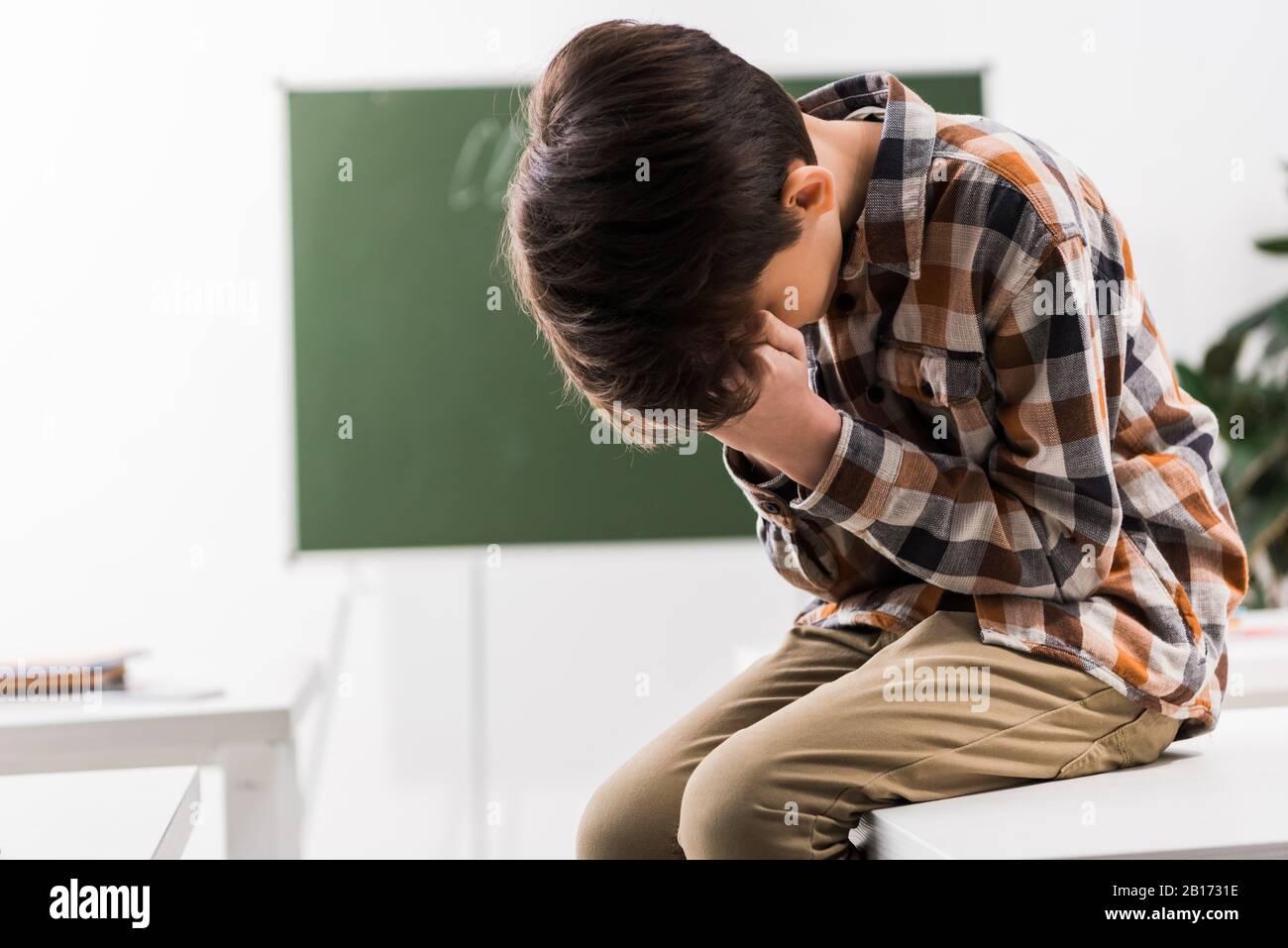 bullied schoolboy covering face while crying in classroom Stock Photo