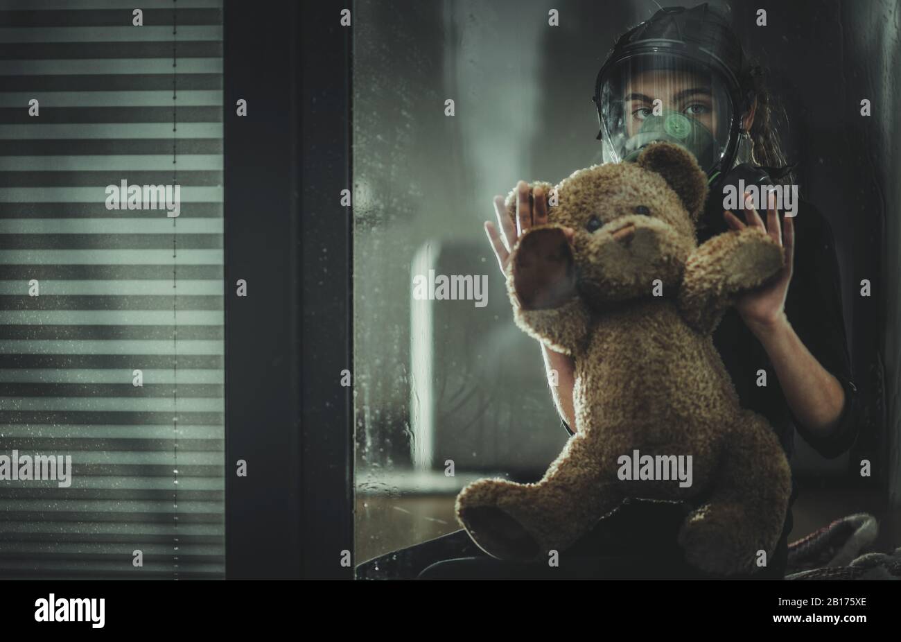 Sad Caucasian Girl Seating with Her Teddy Bear Inside Home with Biochemical Protective Mask and Looking Outside the Window. Dangerous Virus Pandemic C Stock Photo