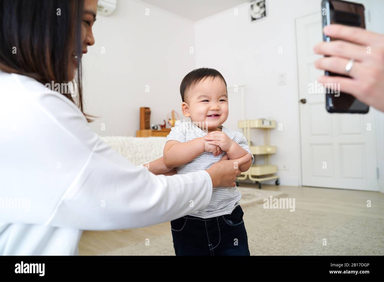 Cute excited little Asian boy enjoying funny video on mobile phone and  laughing while standing in room supported by happy mom Stock Photo - Alamy