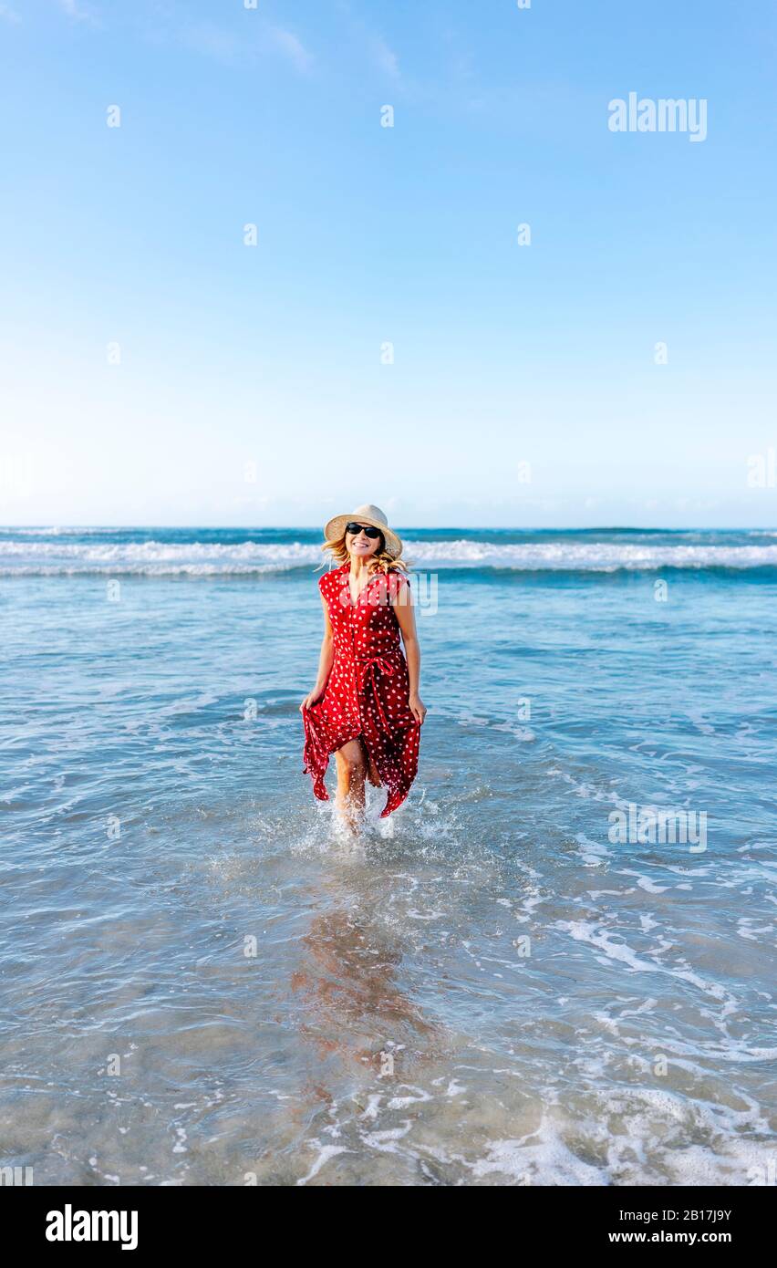 Blond woman wearing red dress and hat at the beach, running in water Stock Photo