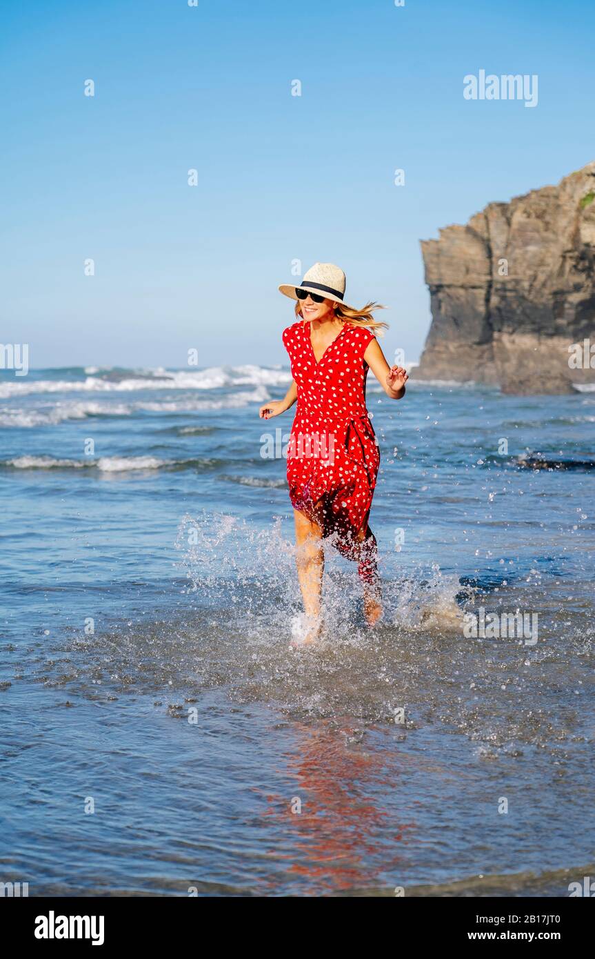 Blond woman wearing red dress and hat and running at the beach, Playa de Las Catedrales, Spain Stock Photo