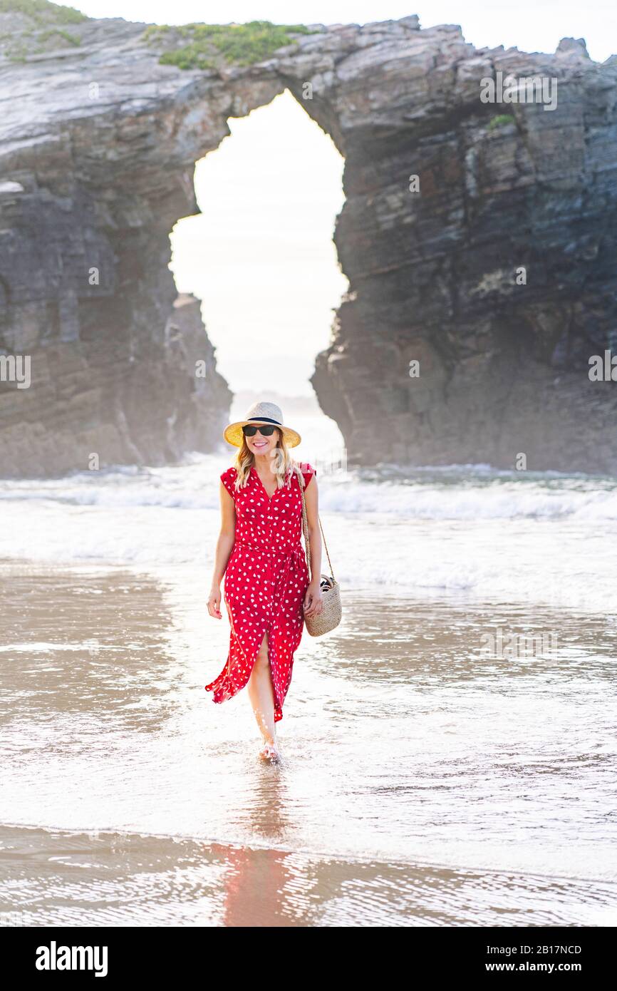 Blond woman wearing red dress and hat and walking at the beach, Natural Arch at Playa de Las Catedrales, Spain Stock Photo