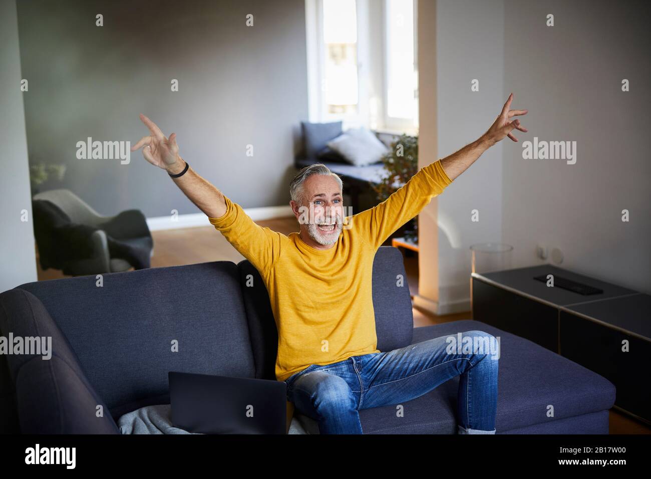 Mature man sitting on couch at home cheering Stock Photo