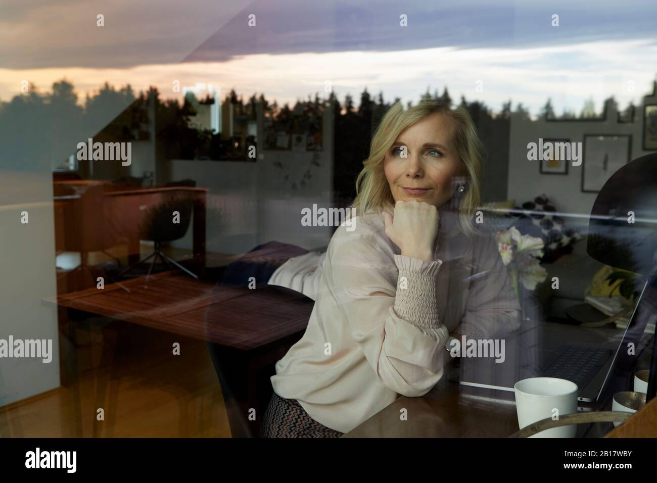 Portrait of blond woman with laptop behind windowpane at home Stock Photo
