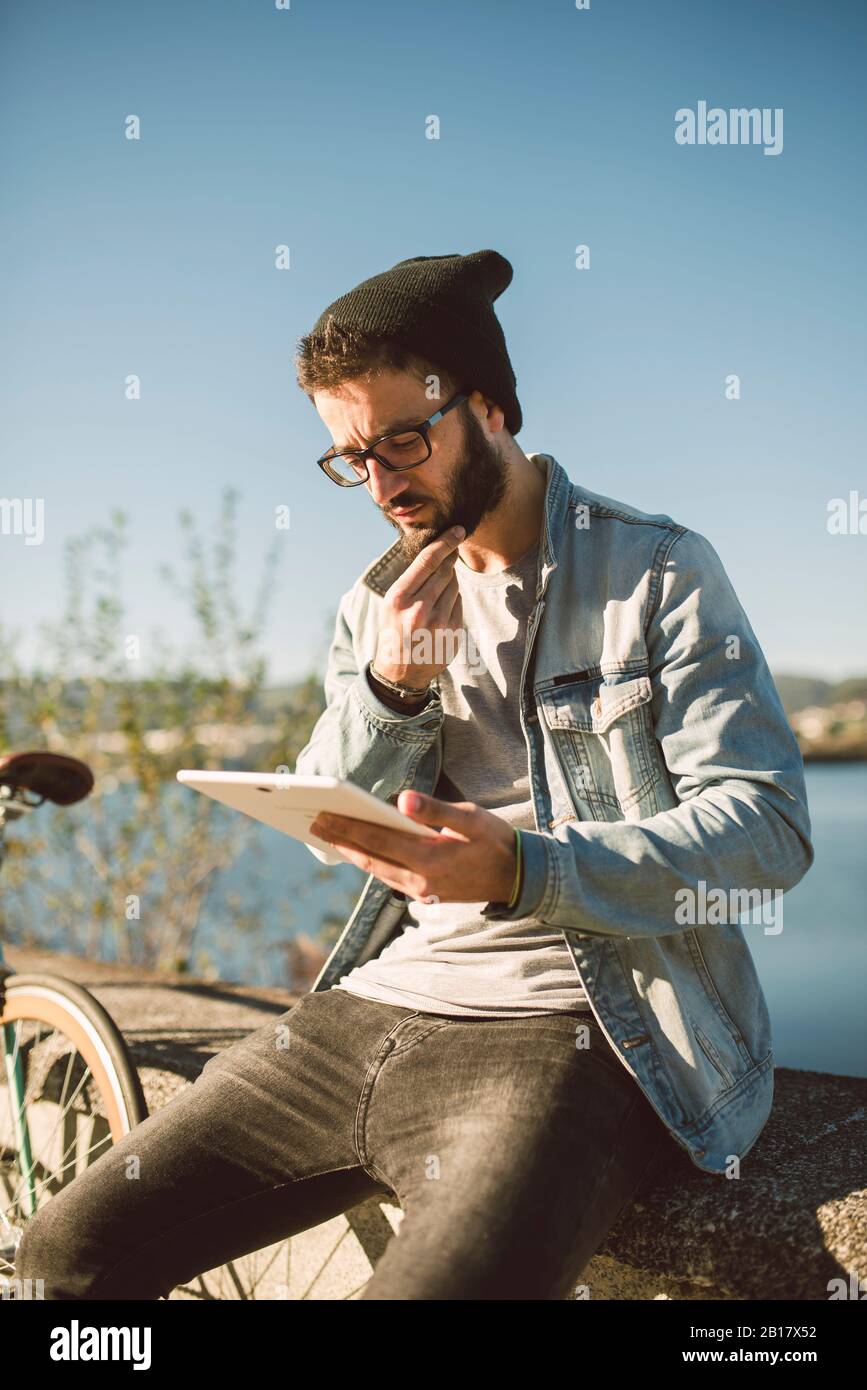 Young man sitting on wal by the sea, using digital tablet Stock Photo