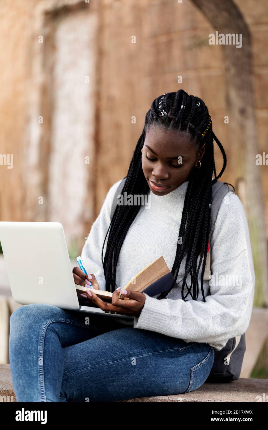 Female student using laptop and taking notes on a bench Stock Photo