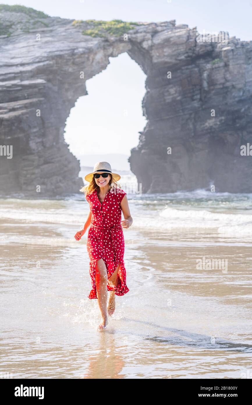 Blond woman wearing red dress and hat and running at the beach, Natural Arch at Playa de Las Catedrales, Spain Stock Photo