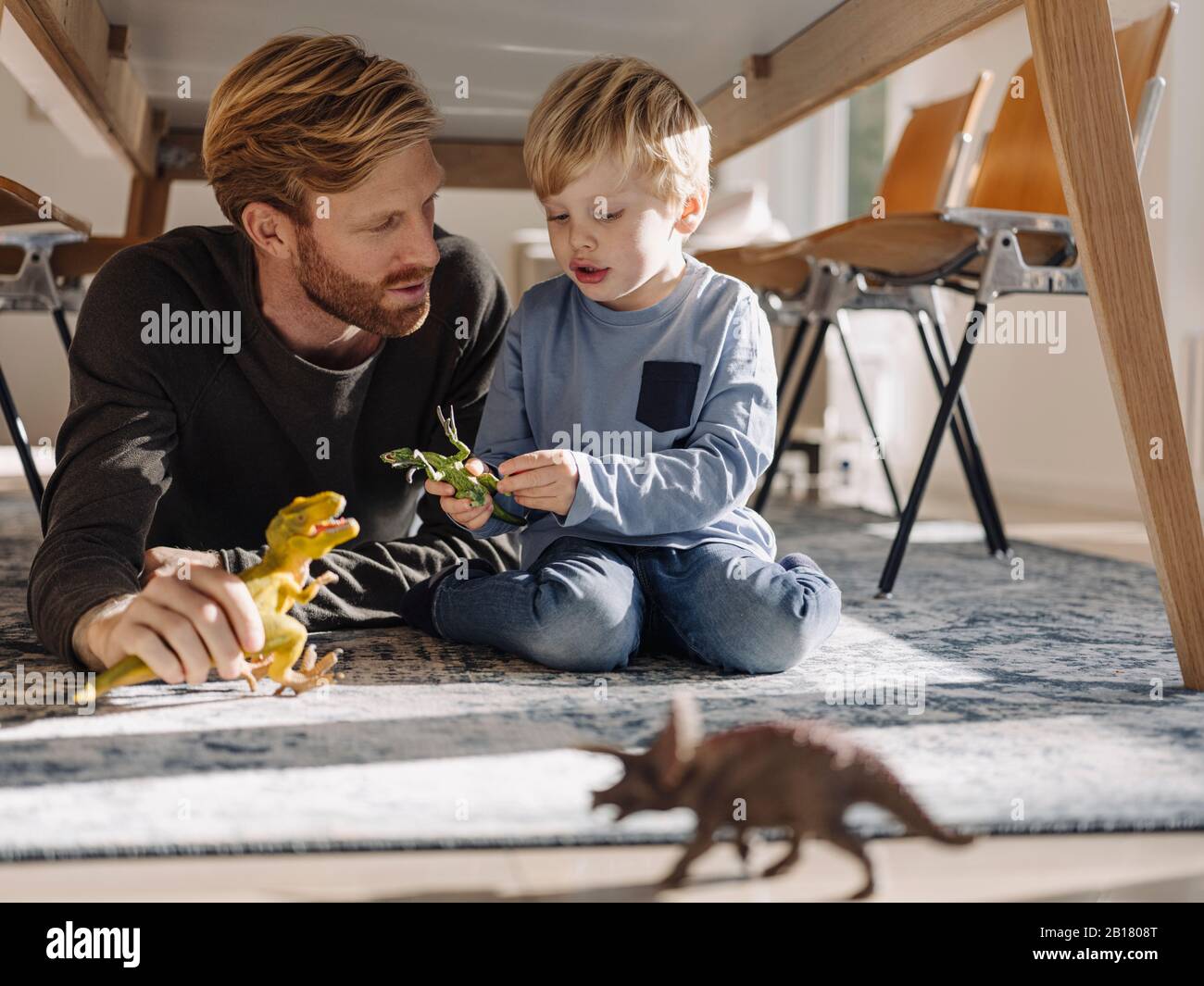 Father and son playing with dinosaur figures under the table at home Stock Photo