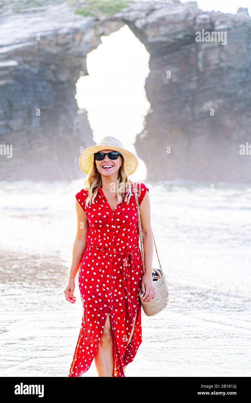 Blond woman wearing red dress and hat and walking along beach, Natural Arch at Playa de Las Catedrales, Spain Stock Photo
