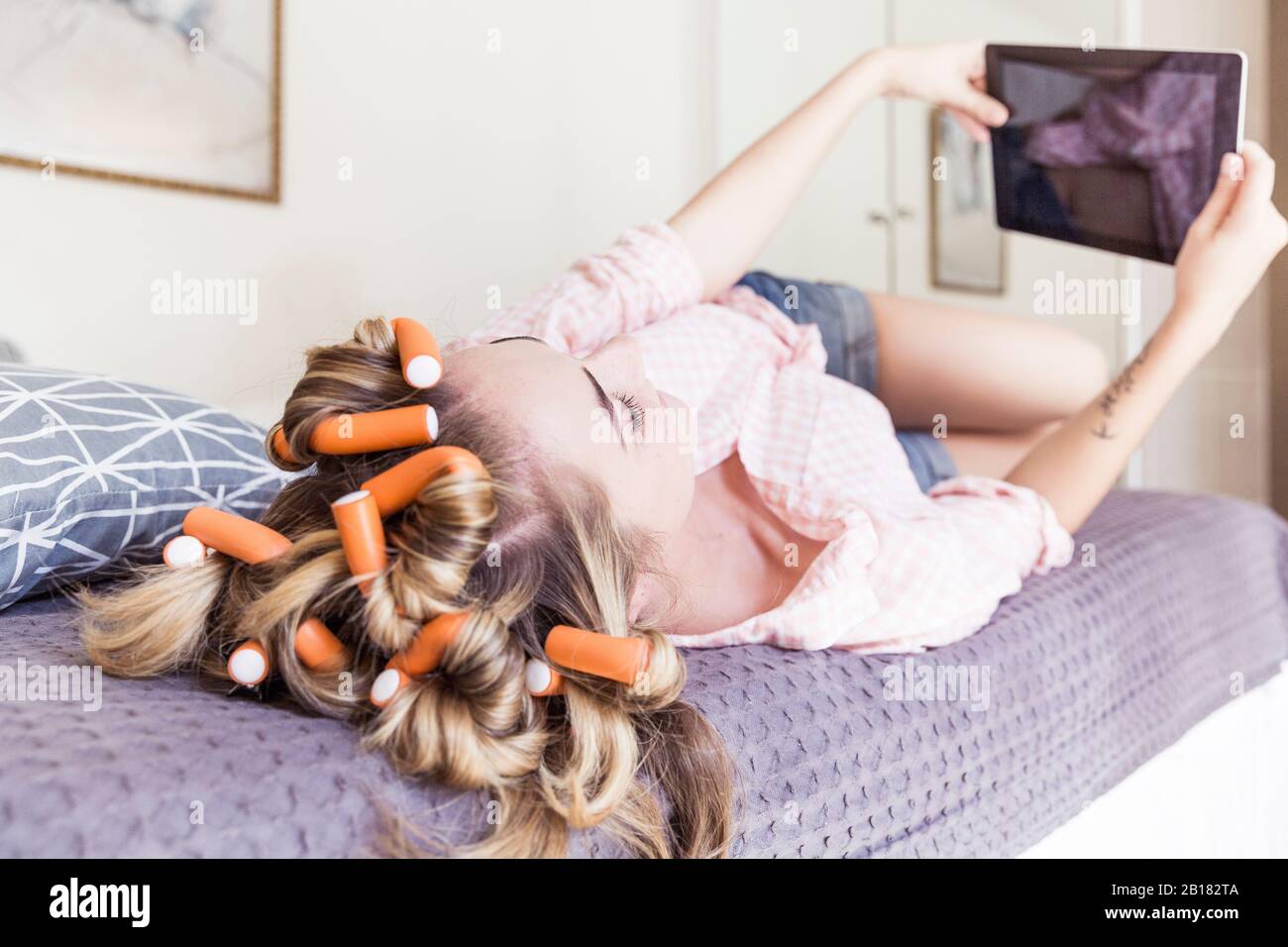 Young woman with curlers in hair lying on bed taking selfie with digital tablet Stock Photo
