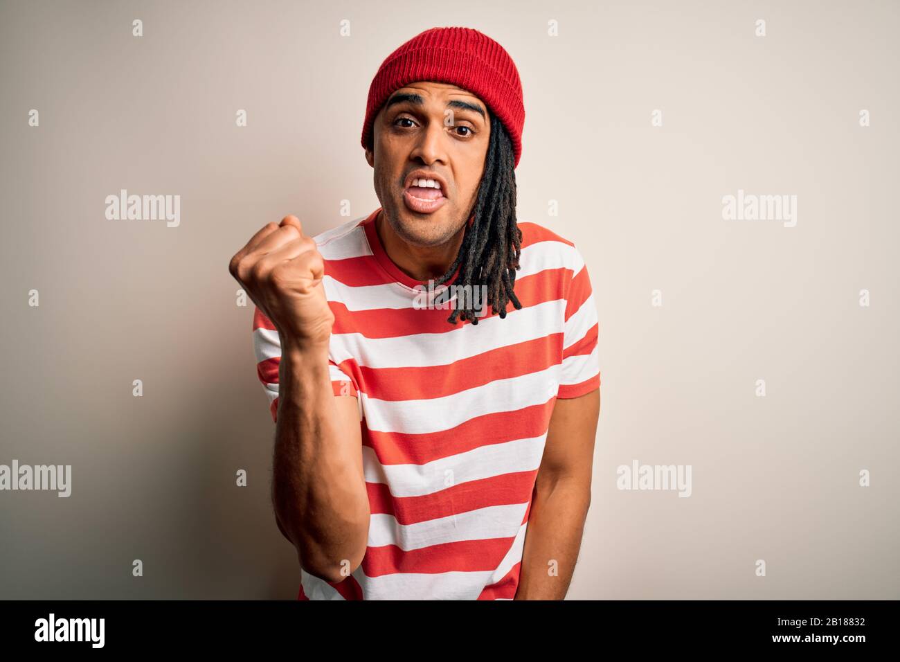 Young handsome african american man with dreadlocks wearing striped t-shirt and wool hat angry and mad raising fist frustrated and furious while shout Stock Photo