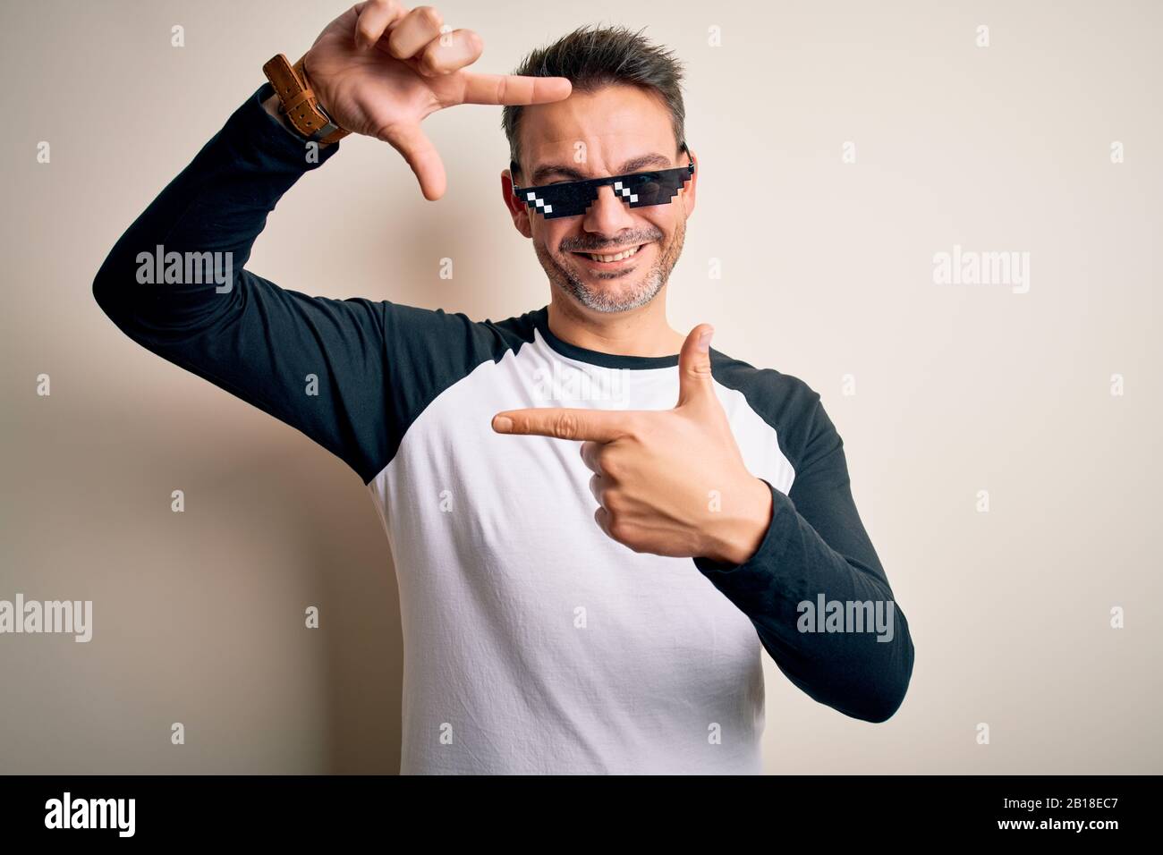 Young handsome man wearing funny thug life sunglasses meme over white background smiling making frame with hands and fingers with happy face. Creativi Stock Photo