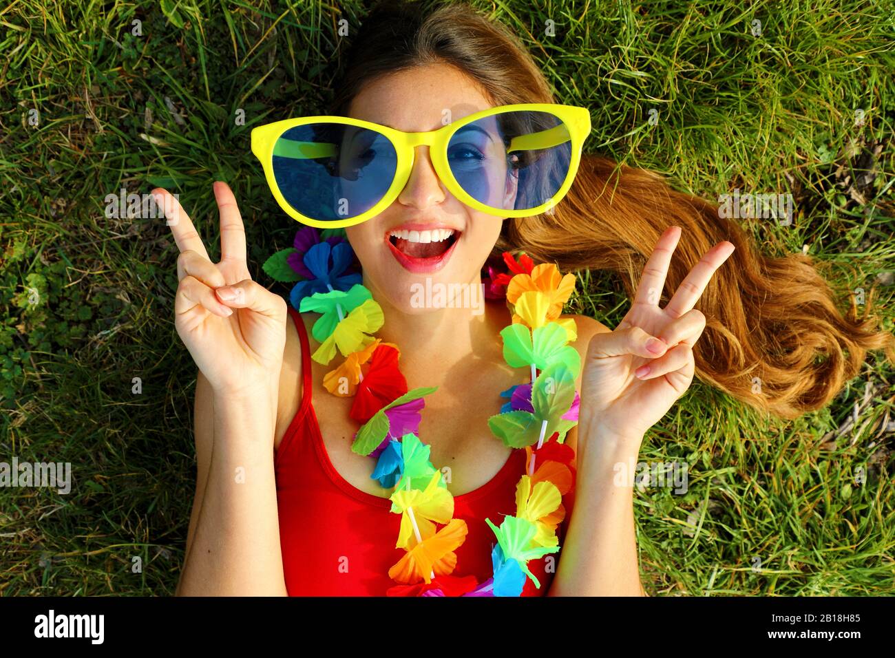 Girl wake up on grass after Carnival party. Young woman with big funny sunglasses and carnival garland lying on grass showing victory V sign with hand Stock Photo