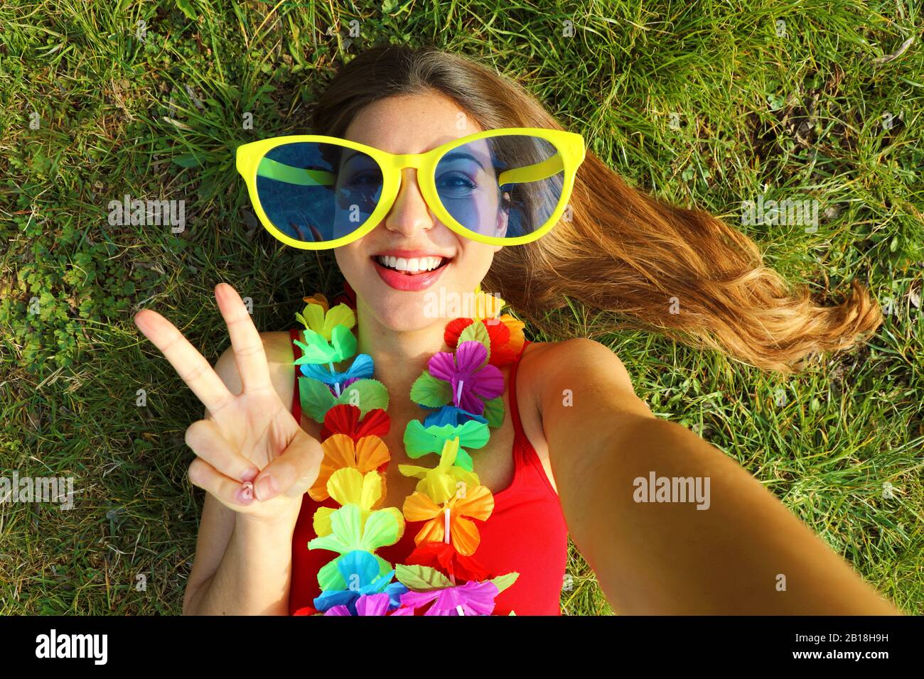 Girl take self portrait after Carnival party. Selfie young woman with big funny sunglasses and carnival garland lying on grass showing victory sign. Stock Photo