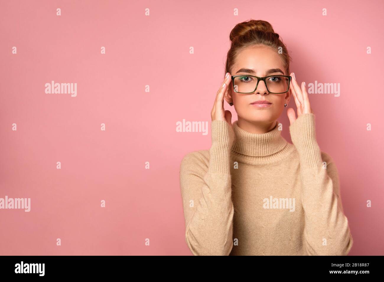 A girl with clean skin and a high bun stands on pink background and looks into the frame, adjusting her glasses with her fingers. Stock Photo