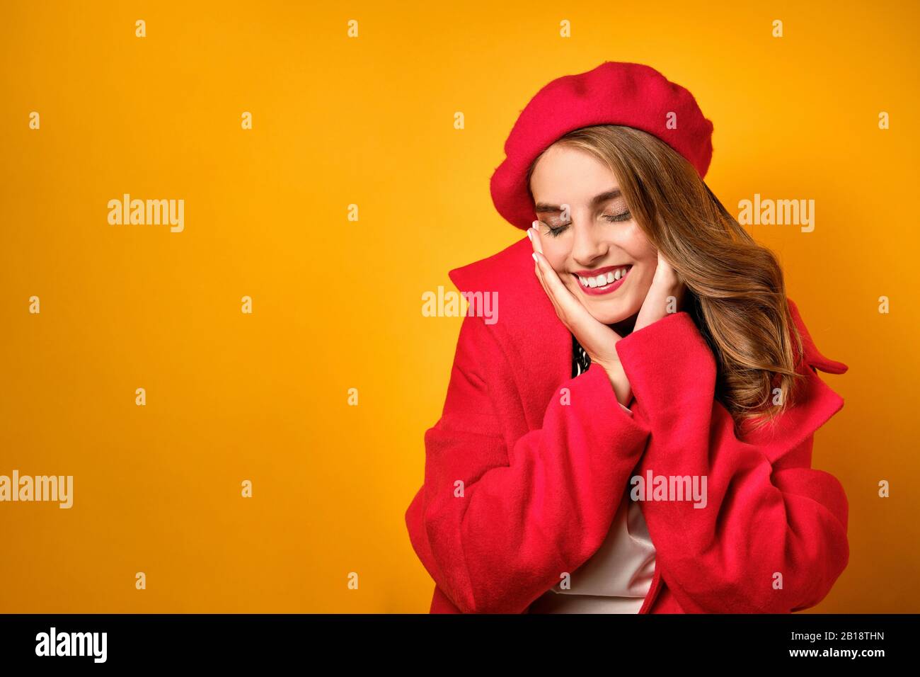 A girl in a red coat and beret is standing on a yellow background and sweetly squinting and smiling, presses her palms to her face Stock Photo