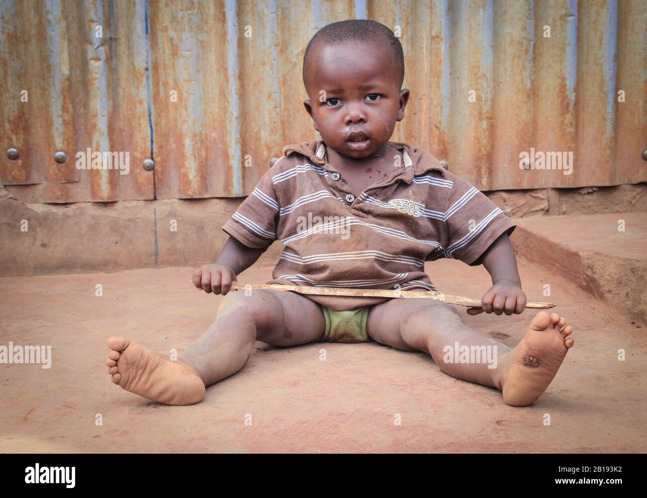 Kibera, Nairobi, Kenya - February 13, 2015: a small dirty black child sits on the floor in the slums and holds a stick in his hands Stock Photo