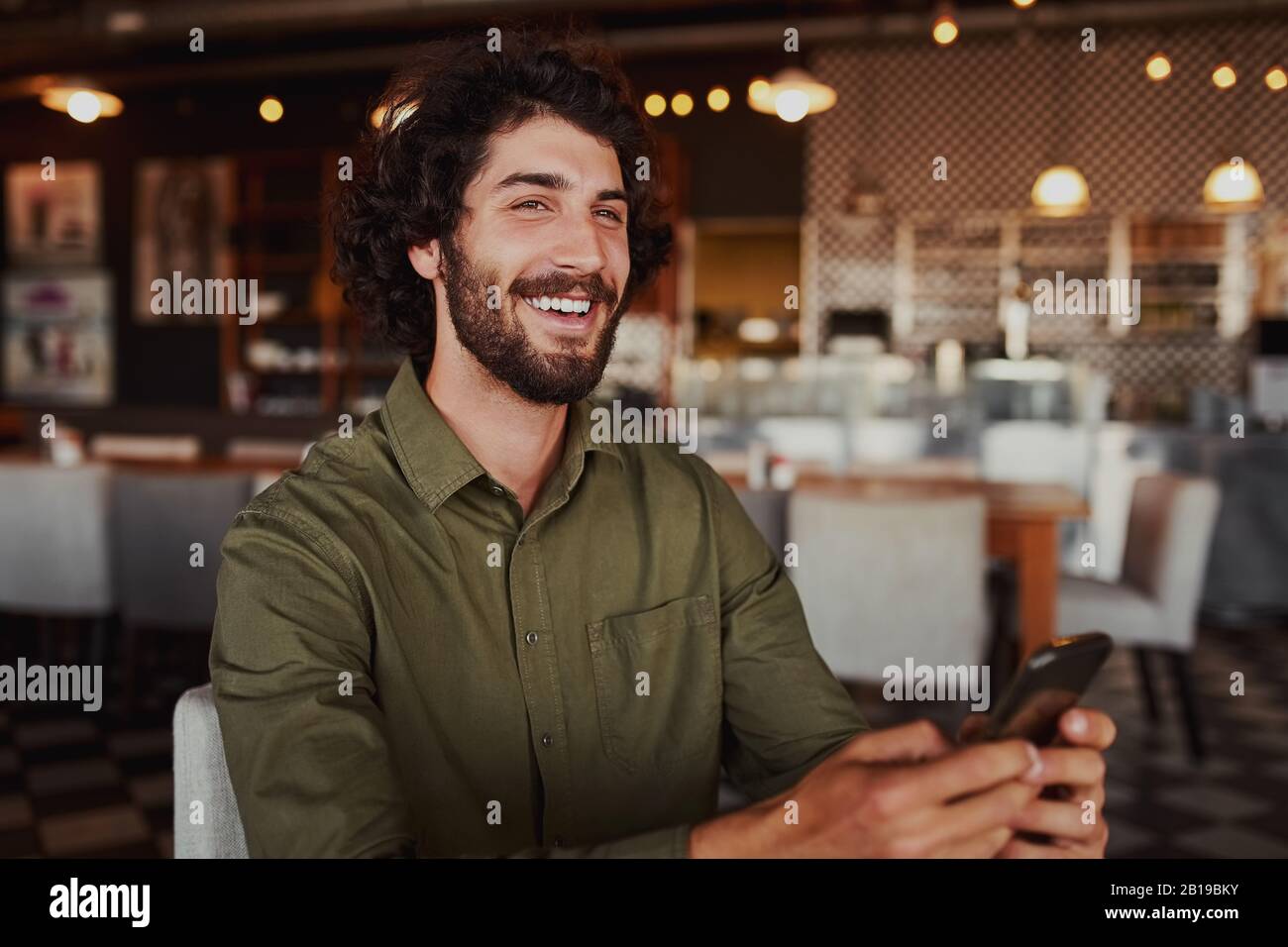 Portrait of handsome young man laughing while reading funny message on phone sitting in modern cafeteria during free time Stock Photo