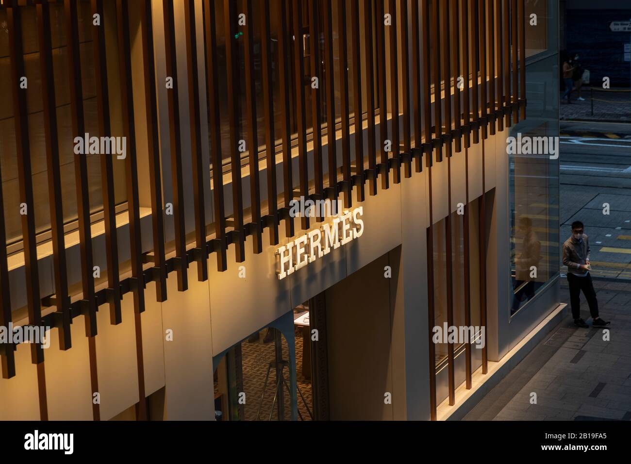 A pedestrian wearing a face mask walks past the Hermes brand store and logo in Hong Kong.The deadly coronavirus (known as COVID-19) has caused most industries, factories and malls in China shut down. More than 50 million people in quarantine. With countries restriction on entries to control the spread of the virus. As well as more than 25,000 flight cancellations worldwide. Many countries cannot get raw material from China. Long dependent on the spending of Chinese buyers remind home. Luxury goods group Herms will provide an update to investors on Wednesday 26 February, 2019. Analysts have war Stock Photo