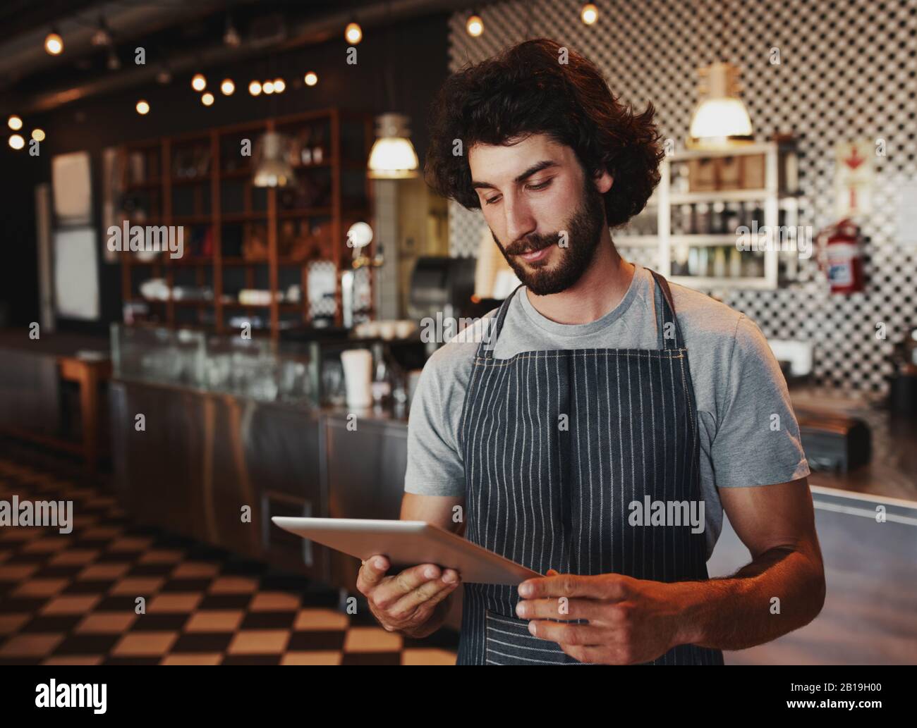 Young cafe owner checking for online order on digital tablet while standing in coffee house wearing black apron Stock Photo