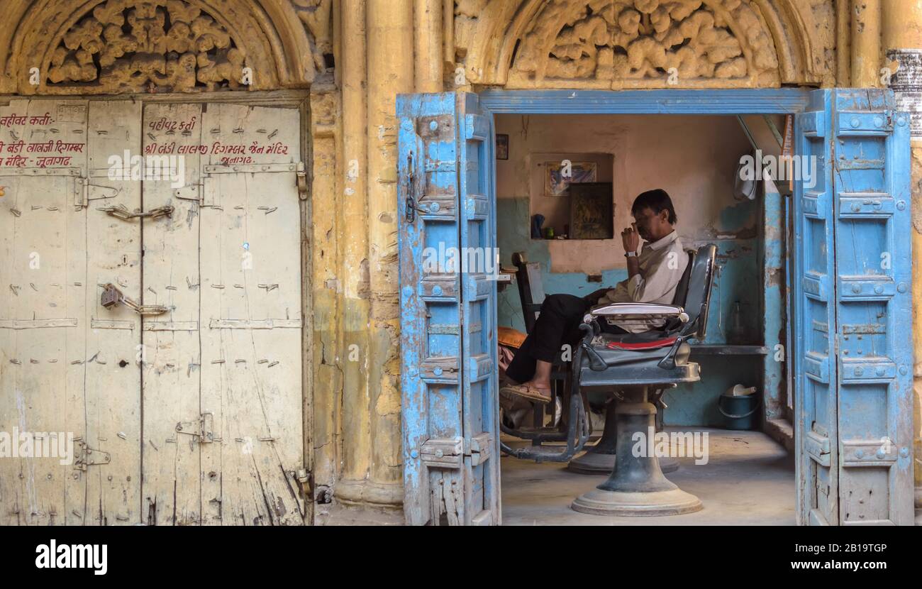 Junagadh, Gujarat, India - December 2018: The quaint blue wooden doors and exterior of a barbershop. A barber sits inside on a chair. Stock Photo