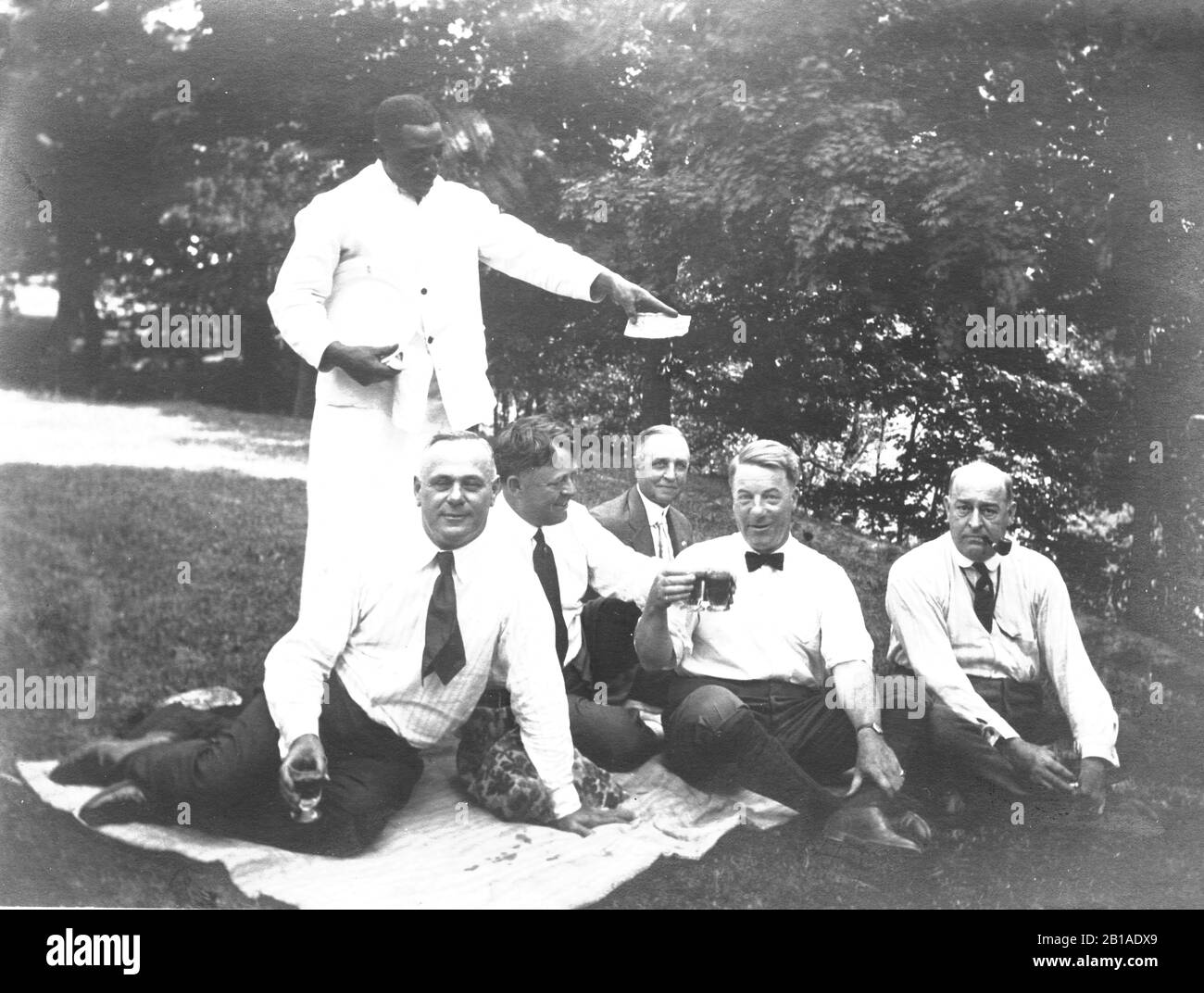One African American waiter/ server holds up a cash tip he received from 5 White businessmen sitting on a picnic blanket. My guess is that the Whites told him to display the cash for the photographer, to show how generous they were. I can't see what amount the cash tip is. The businessmen are enjoying beer, having a good time.  No known location or people.   To see my other African American-related vintage images, Search:  Prestor  vintage  African Stock Photo