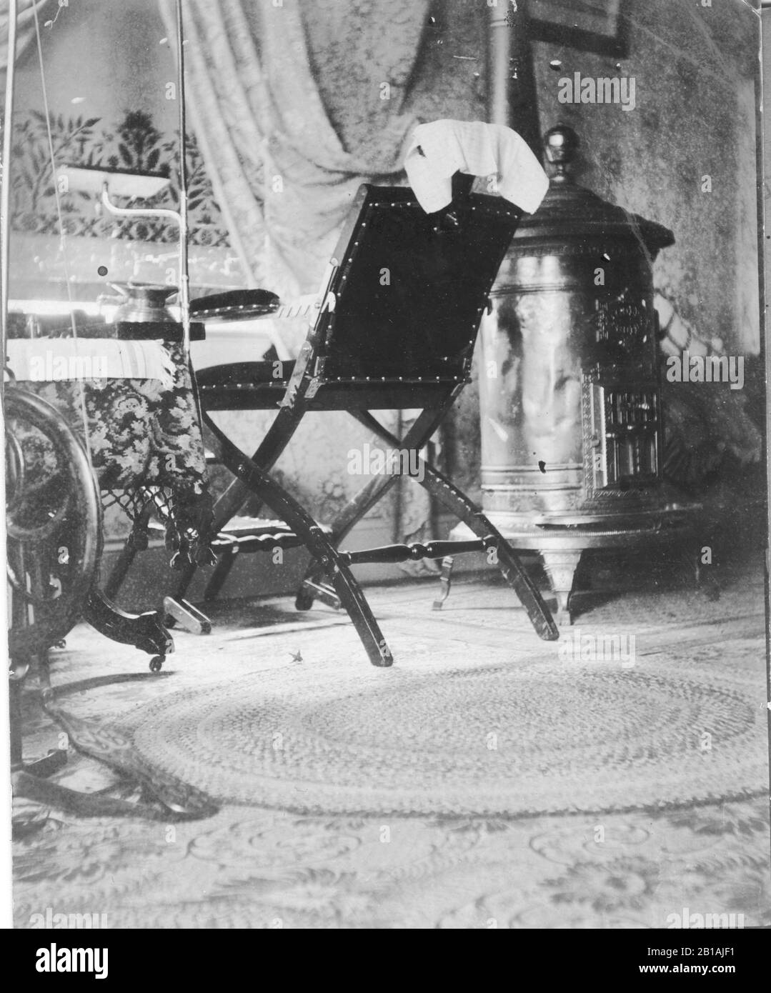 Dental chair, upholstered, c. 1875. In those days, the local dentist was often the barber, too. This seems to be a home practice, not a business in an office. The dental chair has scissors-style folding legs, headrest with a white towel over it, and padded armrests.  At left are a brass spittoon and other dental equipment I cannot identify.  A Potbellied stove is right next to the dental chair. Partially showing at far left are the foot pedal and spinning wheel of a sewing machine.  Unknown location.  To see my other dental-related vintage images, Search:  Prestor  vintage  dental Stock Photo