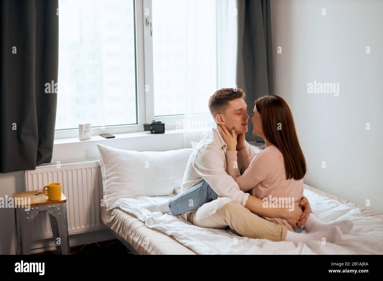 Lovely romantic man and woman going to kiss, look at each other as sit on bed, side view full length photo. tenderness, love Stock Photo