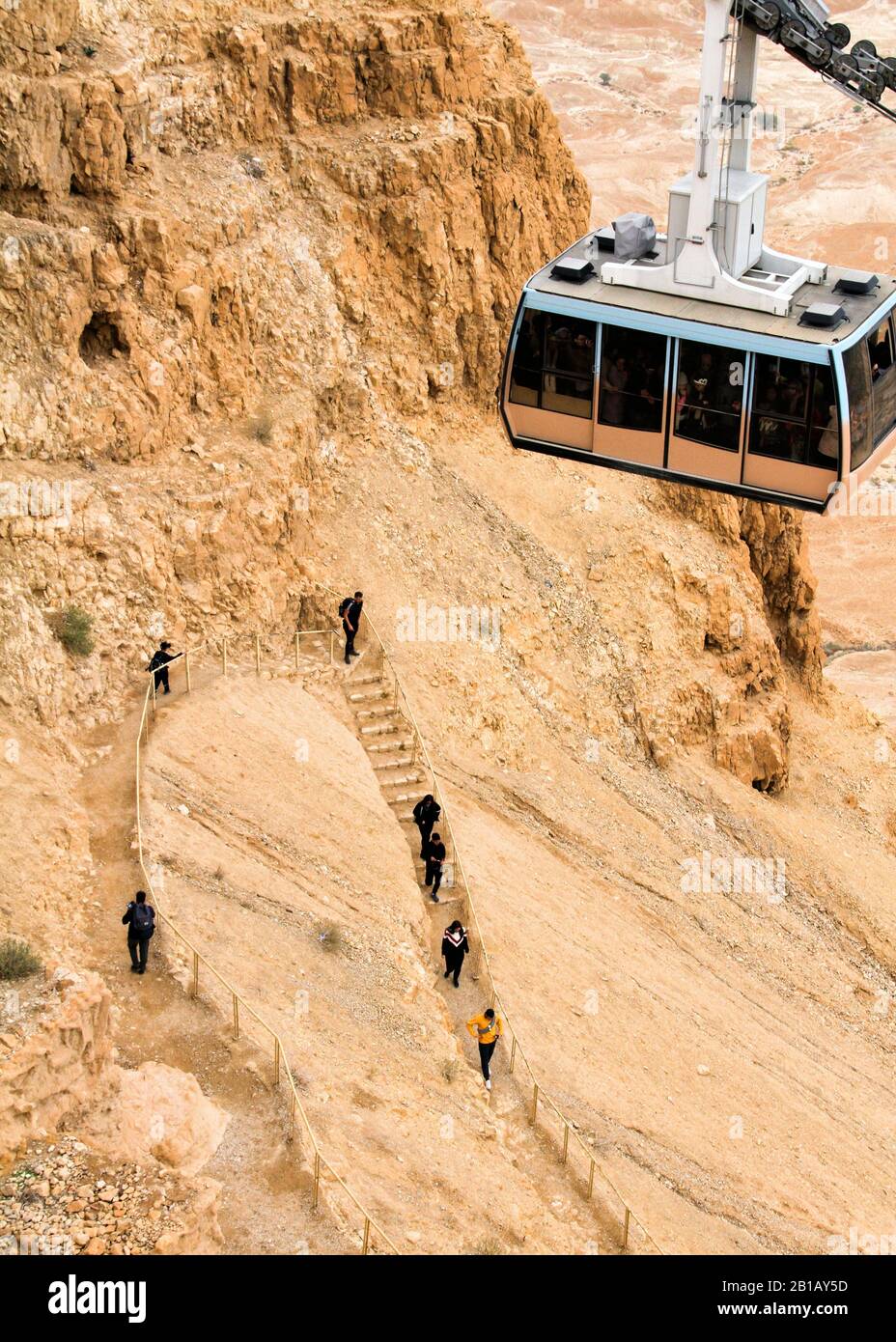 The cable car descends from the top of Masada as people walk down below it. Stock Photo