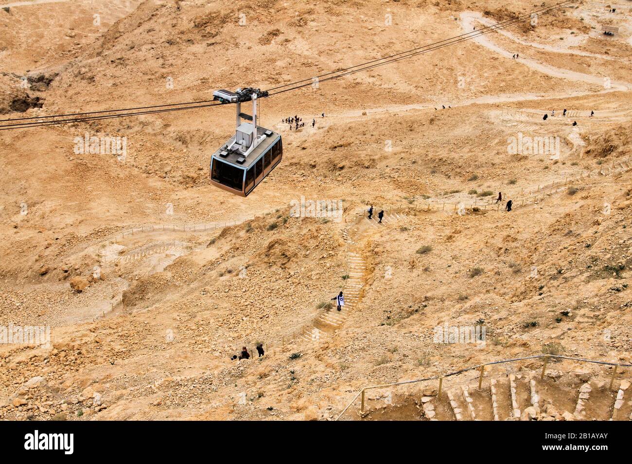 The cable car at Masada National Park in southern Israel descends from the top as people walk down below it. Stock Photo