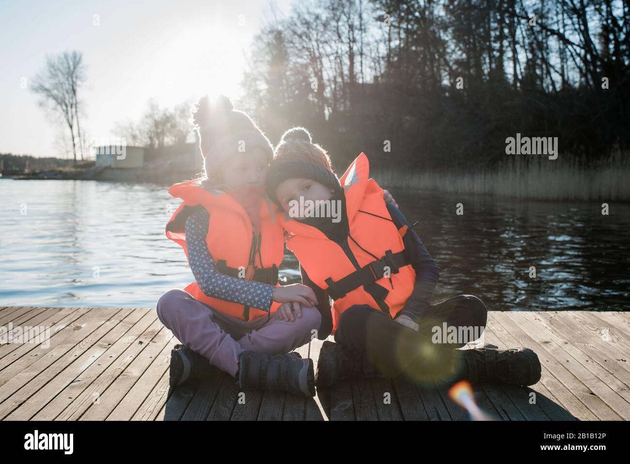 brother and sister sat with life jackets on at the beach hugging Stock Photo