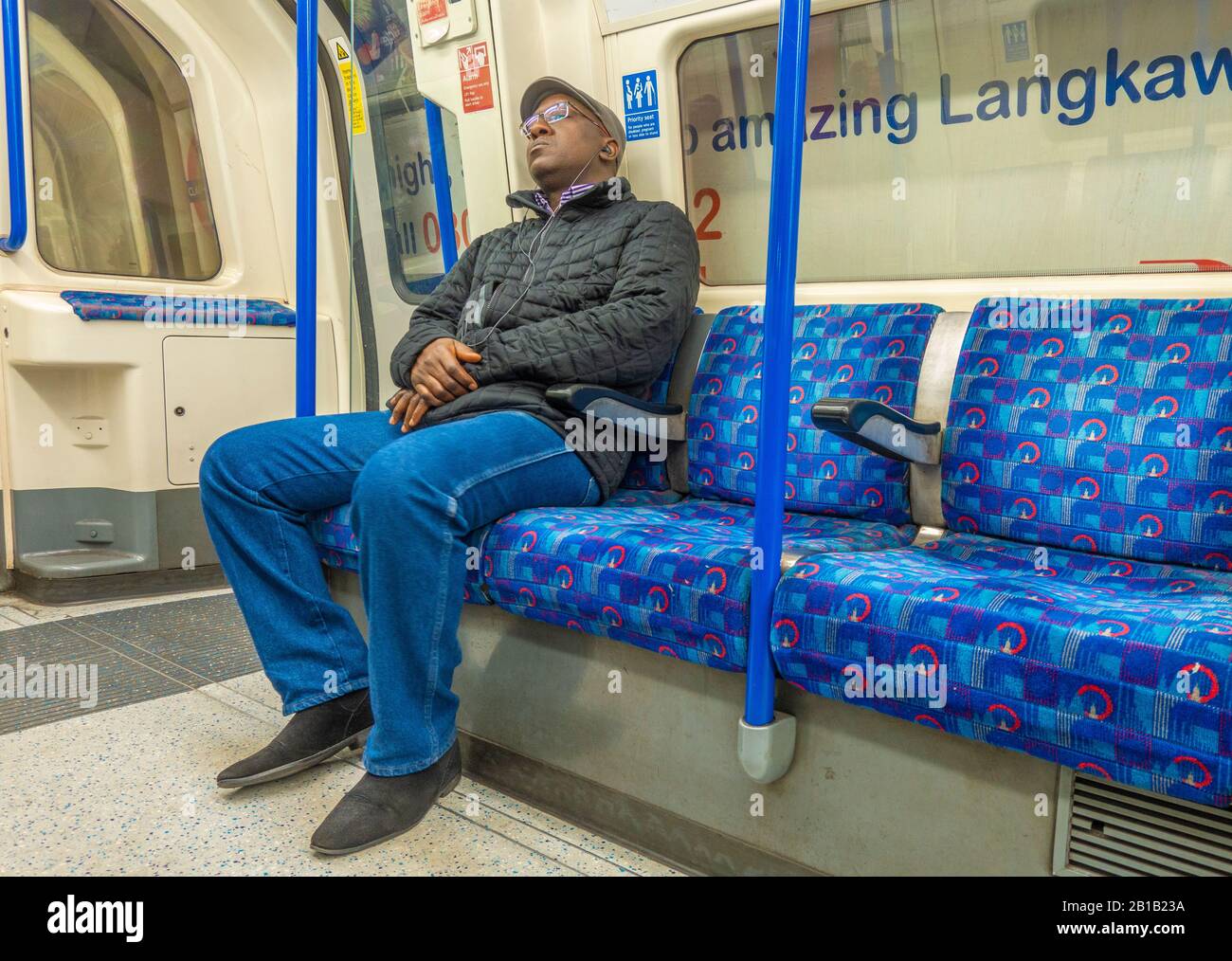 Black man / male seated passenger, dozing on a quiet London Underground tube train carriage, and listening to music through headphones. England, UK. Stock Photo