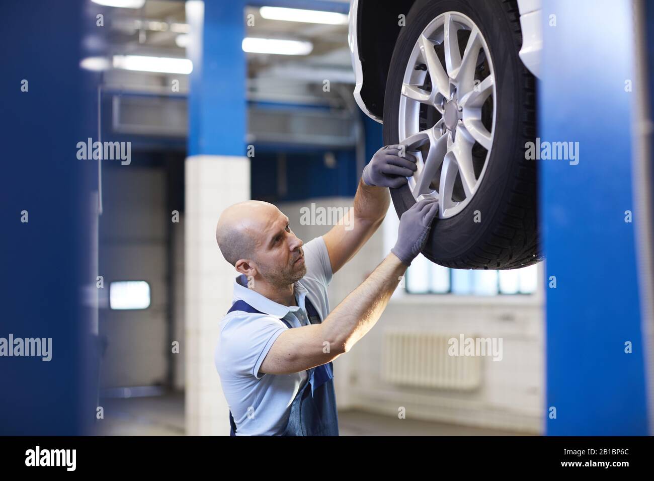 High section portrait of bald car mechanic checking wheels of vehicle on car lift during inspection in garage shop, copy space Stock Photo