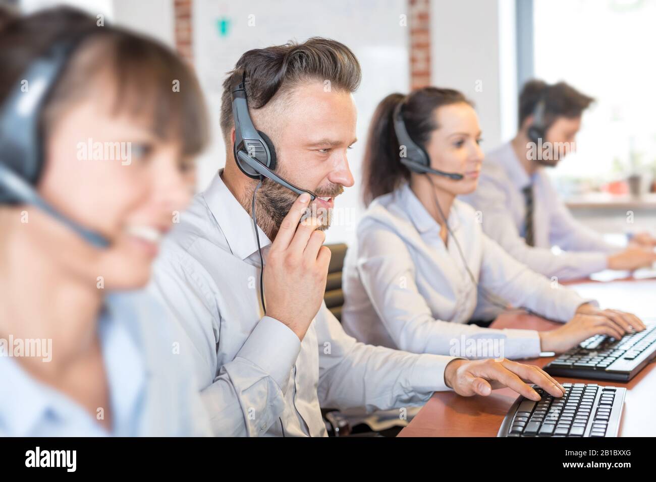Call center worker accompanied by his team. Smiling customer support operator at work. Young employee working with a headset. Stock Photo